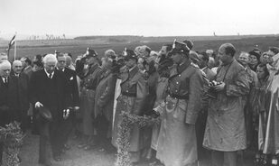 Theodor Heuss schreitet im schwarzen Cut und mit abgesetztem Zylinder der Festansprache entgegen. Die uniformierten Polizeibeamten grüßen militärisch durch Handanlegen an den Rand ihrer Tschakos.