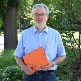 Dr. Norbert Schnitzler steht in einem Park. In der Hand hält er eine Akte.