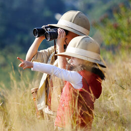 Zwei Kinder stehen im hohen Gras und schauen mit einem Fernglas in die Ferne. Auf den Köpfen tragen sie Safari-Hüte.
