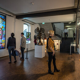 Ein Blick in das Glasmalerei Museum Linnich. Drei Menschen schauen sich Bilder an.