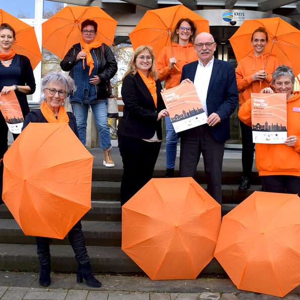 Eine Gruppe Menschen steht mit orangefarbenen Schirmen in den Händen vor dem Kreishaus