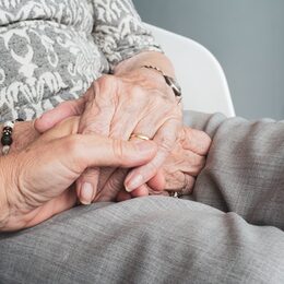 Eine ältere Frau hat ihre Hände im Schoß. Jemand hält ihre Hand.