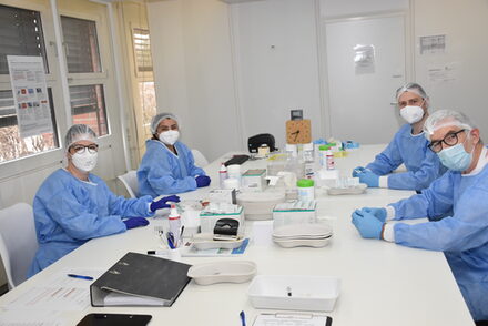 Vier Mitarbeiter in Schutzanzügen sitzen an einem Tisch und bereiten den Impfstoff vor.