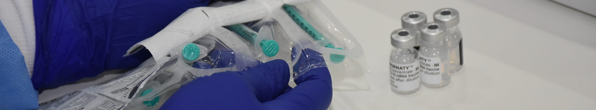 Verpackte Impfspritzen werden ausgepackt von einer Mitarbeiterin in blauen Handschuhen. Nur die Hände sind zu sehen