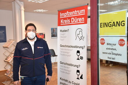 Rolf Corda, Leiter des Impfzentrums steht am Eingang des Impfzentrums vor einer kleinen Leinwand, die die wichtigsten Fragen, die vor dem Betreten zum Gesundheitszustand gestellt werden, aufführt.