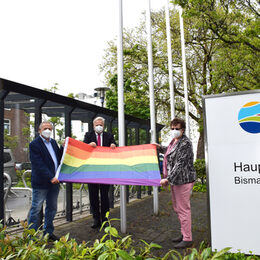LandratWolfgang Spelthahn steht mit zwei Vertretern der Politik vor den Flaggenmasten vor dem Kreishaus und hält die Regenbogenflagge.