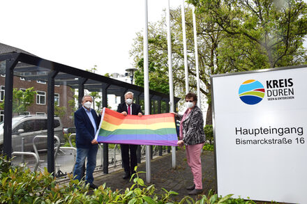 LandratWolfgang Spelthahn steht mit zwei Vertretern der Politik vor den Flaggenmasten vor dem Kreishaus und hält die Regenbogenflagge.