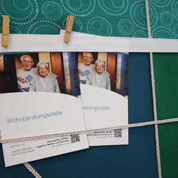 Auf einer Stoff-Pinnwand hängen mit Wäscheklammern befestigt zwei Flyer zur Wohnberatungsstelle, auf denen ein älteres Paar abgebildet ist, die in die Kamera lächeln