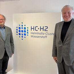 Das Bild zeigt zwei Männer vor einem Schild, auf dem Helmholtz Cluster Wasserstoff steht
