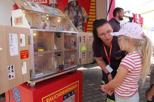 Eine junge Frau erläutert einem Mädchen an einem Modellhaus, wie die Feuerwehr arbeitet.