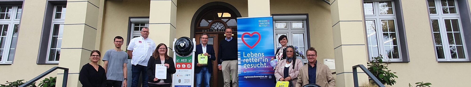 Eine Gruppe Menschen vor dem Rathaus Merzenich. Sie präsentieren Defibrillatoren.
