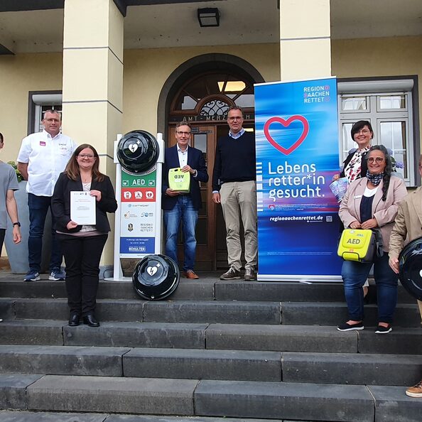 Eine Gruppe Menschen vor dem Rathaus Merzenich. Sie präsentieren Defibrillatoren.