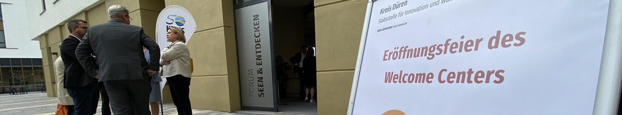 Das Bild zeigt ein Hinweisschild und eine Gruppe von Menschen vor dem neuen Welcome Center