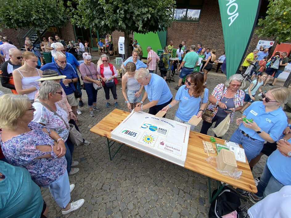 Das Foto zeigt den Landrat, wie eine große Torte anschneidet. Um den Tisch stehen mehrere Menschen.