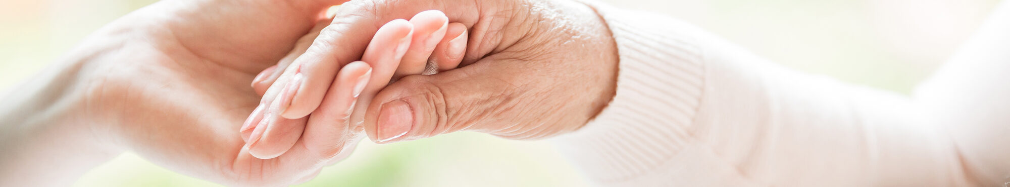 Close-up of tender gesture between two generations. Young woman holding hands with a senior lady. Blurred background. Panorama.