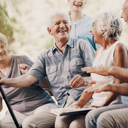 Senioren lachen gemeinsam im Garten und eine Pflegerin ist im Hintergrund zu sehen