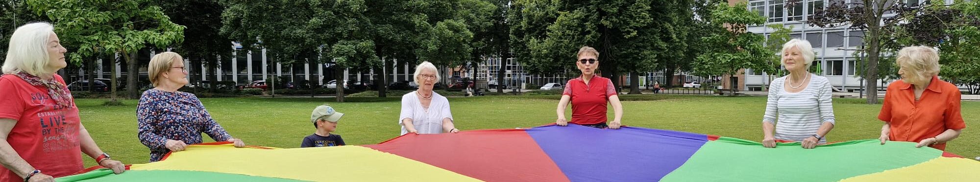 Senioren und Kinder stehen zusammen im Kreis und schwingen ein großes Tuch in bunten Farben.