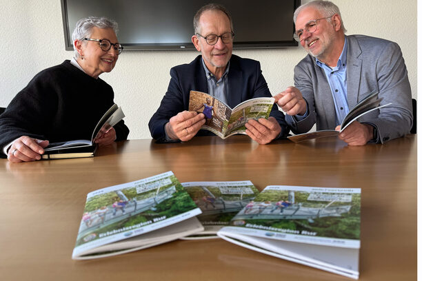 Das Bild zeigt zwei Männer und eine Frau an einem Tisch; die blättern in der Broschüre